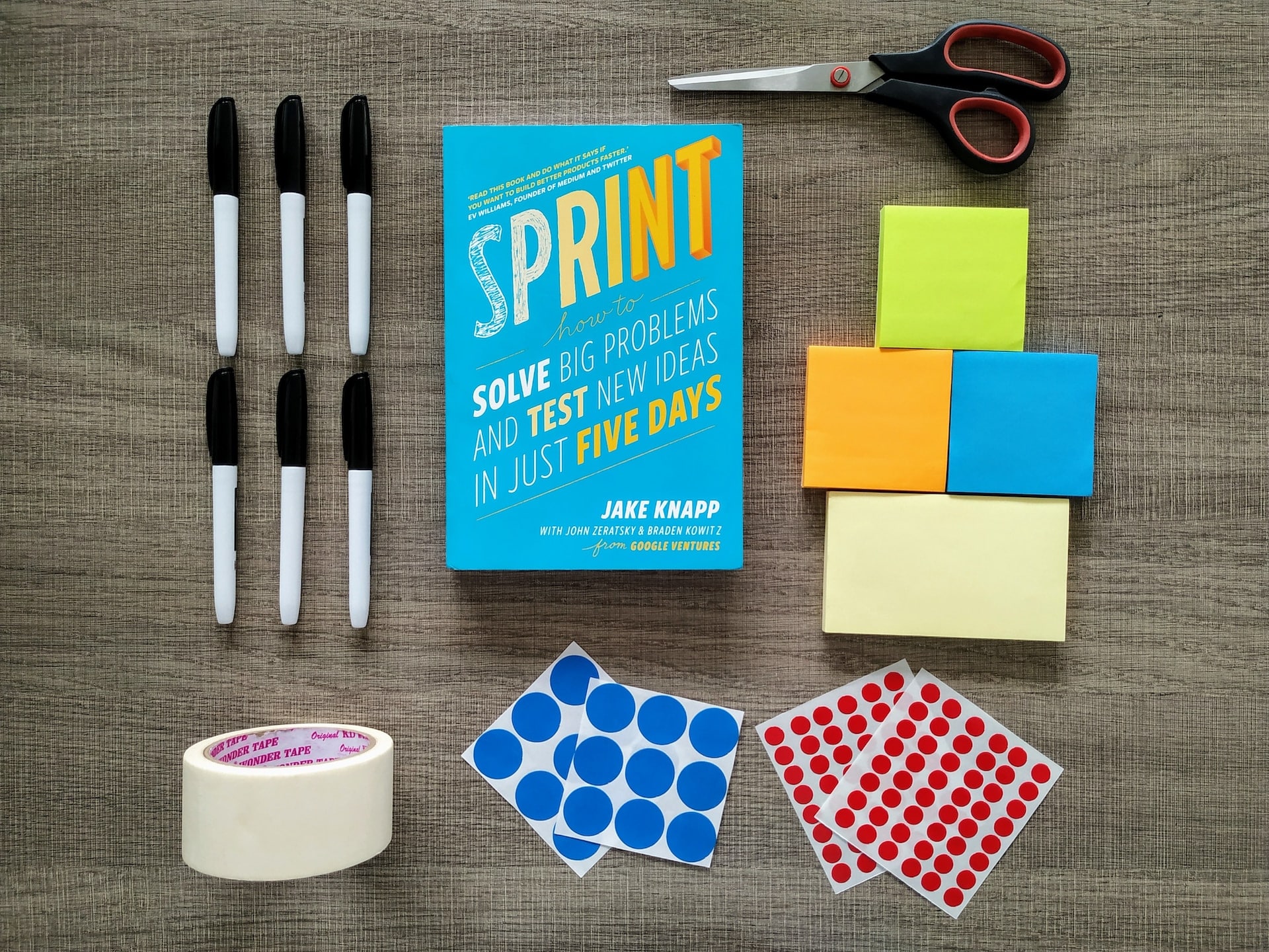 Foto of a desk showing a printed copy of the book "Sprint", a scissors, 6 pens, masking tape, voting dots and four packs of sticky notes.